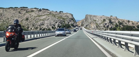 Eine Straße auf der Insel Krk mit viel Verkehr, darunter ein Motorrad und mehrere Autos, umgeben von felsigen Hügeln.